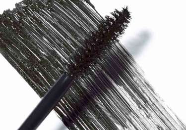 A black mascara brush smears dark mascara across a white surface, showcasing the bristles and the texture of the applied product.