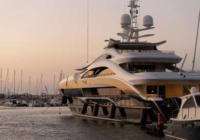 A large luxury yacht is docked at a marina during sunset, surrounded by smaller boats and masts, with calm water reflecting the evening light.