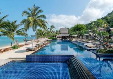 Hotel pool next to beach with deck chairs and palm trees.
