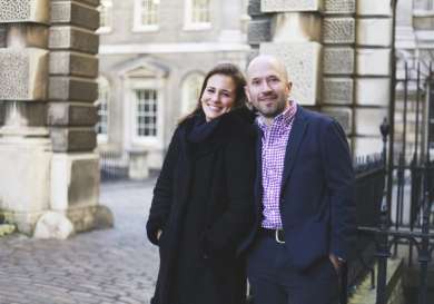 Two people stand smiling in a cobblestone courtyard, leaning casually against a black wrought iron railing, surrounded by historic stone buildings with large windows.