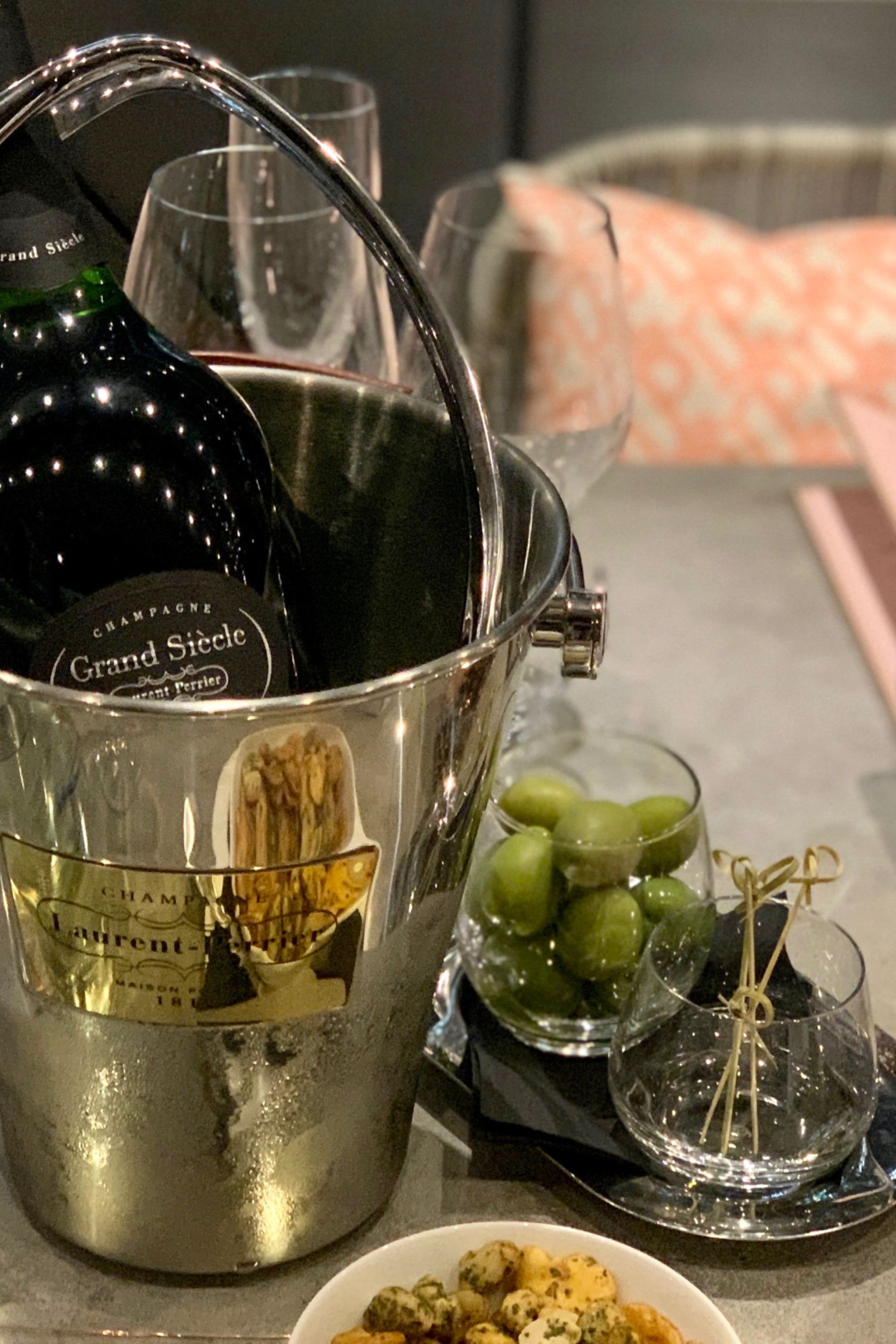 A bottle of Grand Siècle champagne chills in an ice bucket on a dining table, accompanied by a pink Laurent-Perrier bottle, green olives, condiments, and folded napkins in a cozy setting