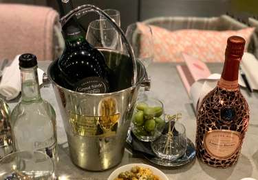 A bottle of Grand Siècle champagne chills in an ice bucket on a dining table, accompanied by a pink Laurent-Perrier bottle, green olives, condiments, and folded napkins in a cozy setting.