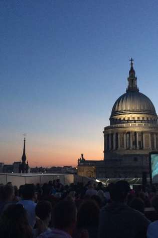 A crowd watches an outdoor movie on a large screen at dusk, with a historic domed building and city skyline in the background under a clear evening sky.