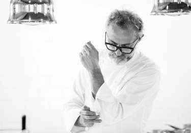 A man wearing glasses and a chef's coat adjusts his sleeve.