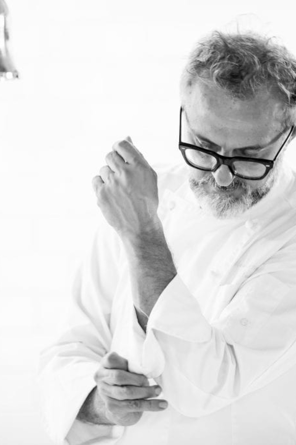 A man wearing glasses and a chef's coat adjusts his sleeve