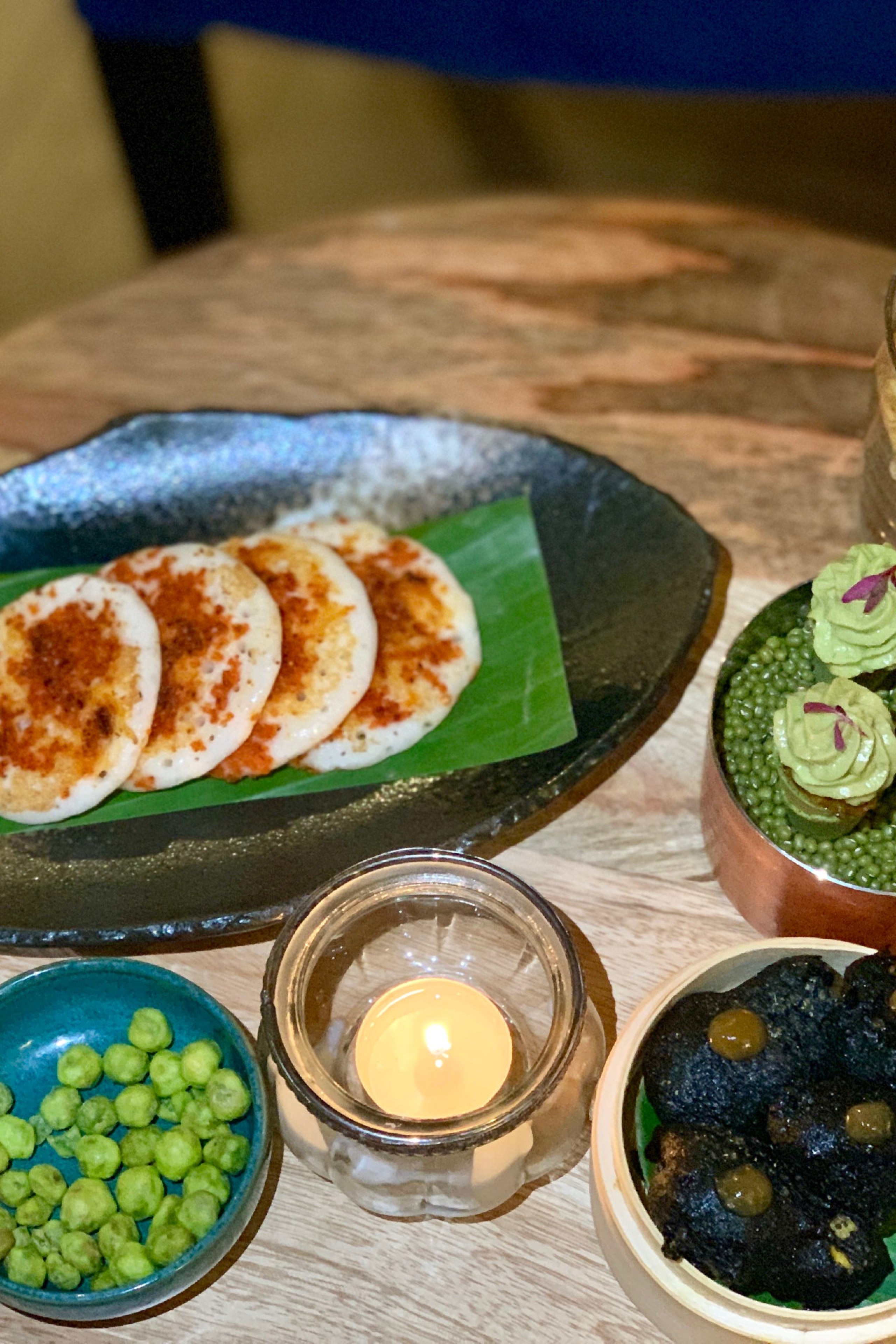 A table displays an array of gourmet appetizers: multiple small pancakes with spices, green peas, crispy snacks in jars, avocado-topped bites, and dark chips