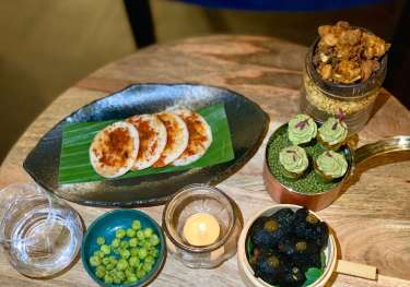 A table displays an array of gourmet appetizers: multiple small pancakes with spices, green peas, crispy snacks in jars, avocado-topped bites, and dark chips.