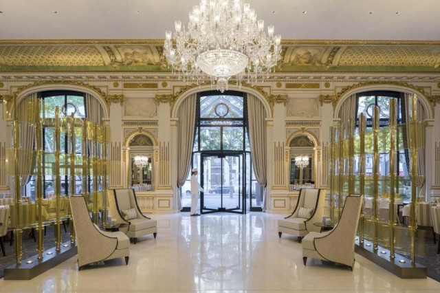 A grand chandelier hangs in an ornate hotel lobby with high ceilings, elegant seating, and floor-to-ceiling windows that offer a view of tree-lined streets