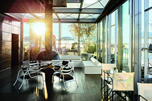 Chairs and tables are arranged indoors, while sunlight streams through large glass windows, highlighting a modern, spacious lounge area