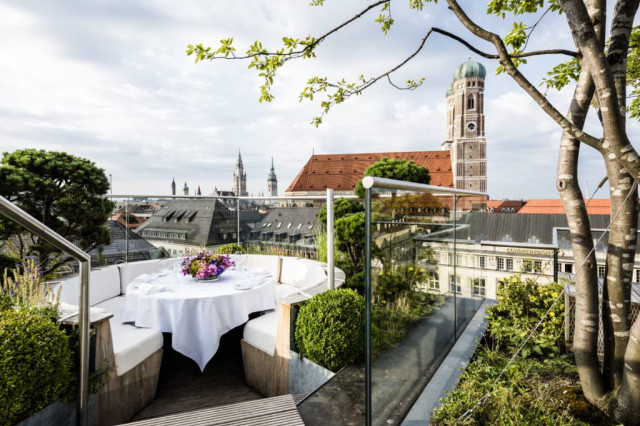 A round table with a white tablecloth and flower centerpiece is set on an outdoor rooftop terrace, surrounded by greenery, overlooking a cityscape with prominent church towers