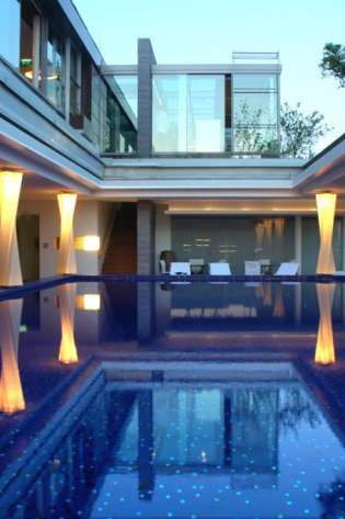 A luxurious indoor pool reflects illuminated columns surrounded by modern architecture, with large glass windows and seating areas on either side, providing a serene and elegant ambiance.