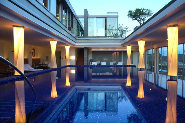 A luxurious indoor pool reflects illuminated columns surrounded by modern architecture, with large glass windows and seating areas on either side, providing a serene and elegant ambiance