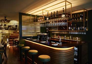 A polished wooden bar counter is lined with stools, displaying wine glasses and bottles overhead.