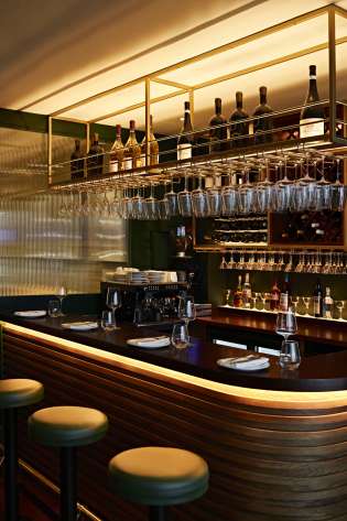 A polished wooden bar counter is lined with stools, displaying wine glasses and bottles overhead.