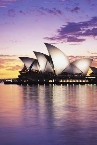 Iconic white sails of the Sydney Opera House are illuminated against a colorful sunset, reflecting on the calm harbor waters, creating a serene and picturesque scene.