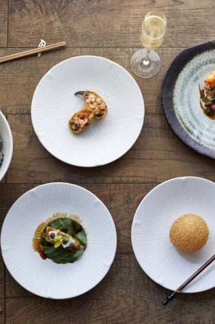 Five elegantly plated dishes rest on a wooden table, accompanied by chopsticks, glasses of champagne, and a small bowl of sauce, creating a sophisticated dining setting.