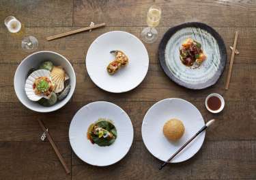 Five elegantly plated dishes rest on a wooden table, accompanied by chopsticks, glasses of champagne, and a small bowl of sauce, creating a sophisticated dining setting.