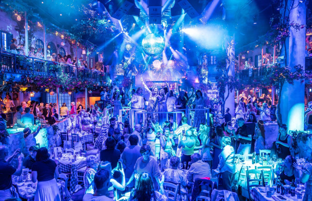 People are dancing and celebrating amid vibrant blue lights under a disco ball, surrounded by tables and balconies adorned with flowers and foliage, suggesting a lively party atmosphere.