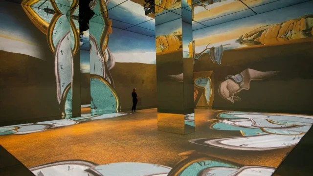Digital art installation projects surreal, melting clocks on mirrored walls and floors, reflecting a vast desert landscape. A solitary person stands observing, adding scale and depth to the scene.