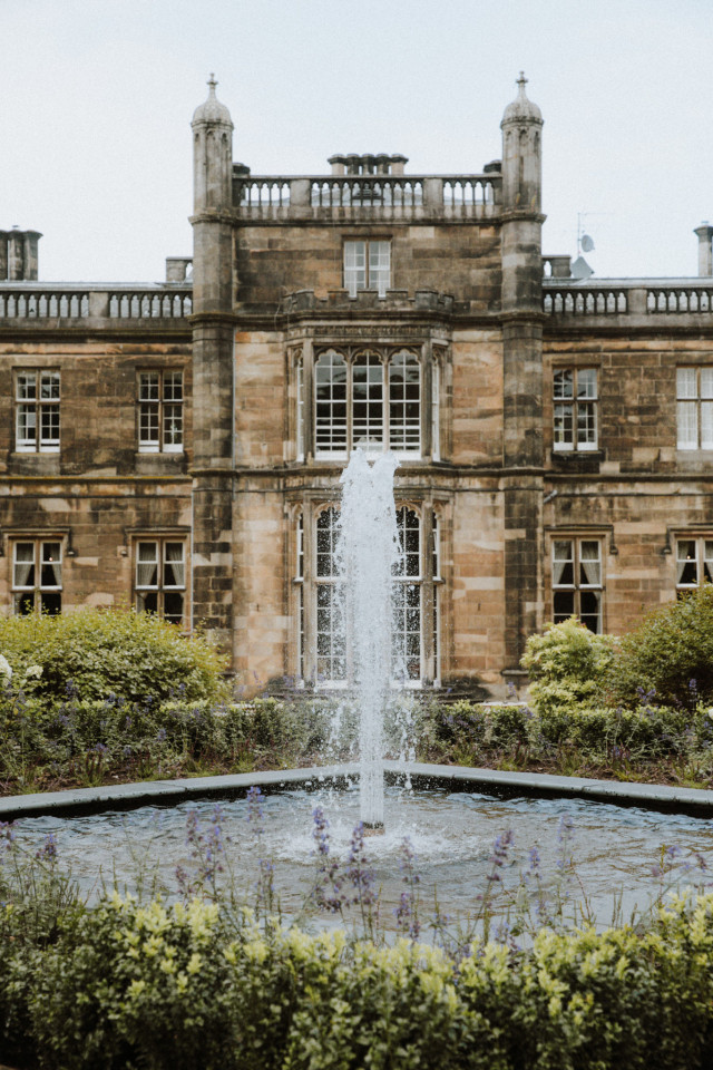A water fountain sprays upward in a landscaped garden, surrounded by shrubs. Behind it stands a large, historic building with stone walls and multiple windows.