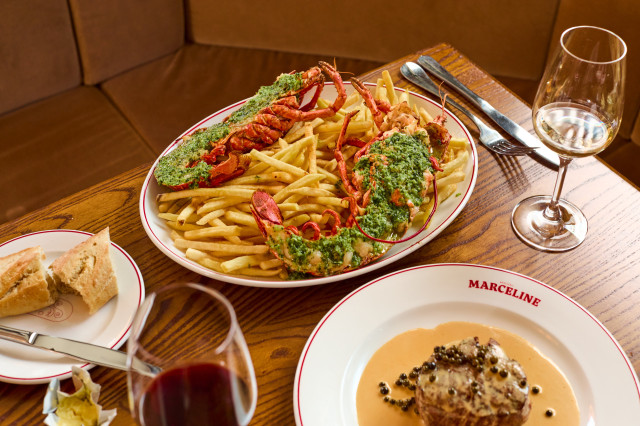 A plate of lobster with herb topping rests on fries, accompanied by steak in sauce. Surrounded by red wine, white wine, bread, and labeled 