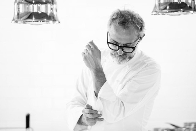 A man wearing glasses and a chef's coat adjusts his sleeve