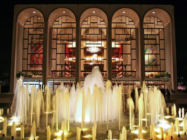 A large fountain cascades in front of an illuminated building with arched windows and colorful interior artwork