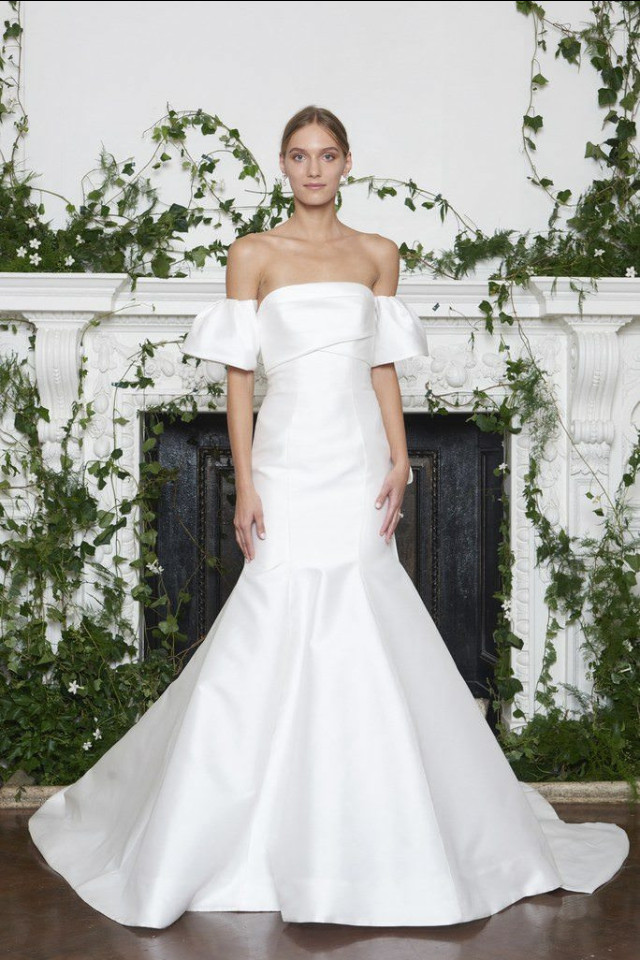 A person stands wearing an off-shoulder, fitted white wedding gown; lush green ivy decorates the fireplace backdrop, creating a classic, elegant setting