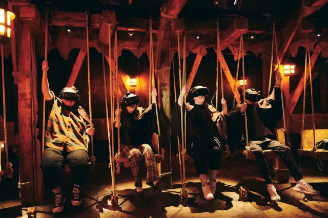 Four people sit on swings wearing virtual reality headsets, immersed in a dimly lit, rustic wooden environment with warm, lantern-like lighting.
