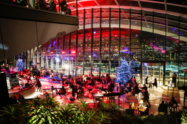 Guests are dining and socializing in a stylish, spacious, glass-walled venue. Festive lighting and a Christmas tree add seasonal decor, with a cityscape view in the background.