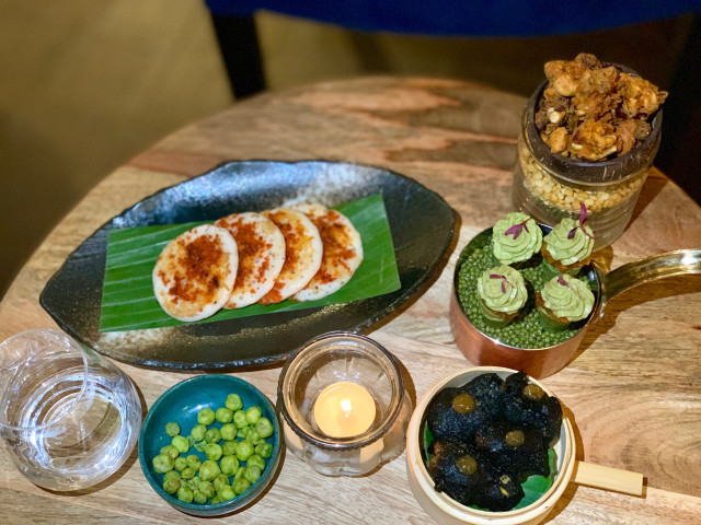 A table displays an array of gourmet appetizers: multiple small pancakes with spices, green peas, crispy snacks in jars, avocado-topped bites, and dark chips