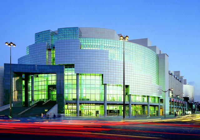 A modern, glass-walled building glows with green lighting as cars streak past in long exposure at twilight, set against a clear blue sky
