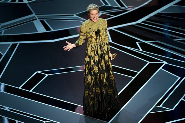 A person in an ornate, long dress stands smiling, gesturing with one hand, holding an award in the other, on a geometrically patterned stage under bright lights