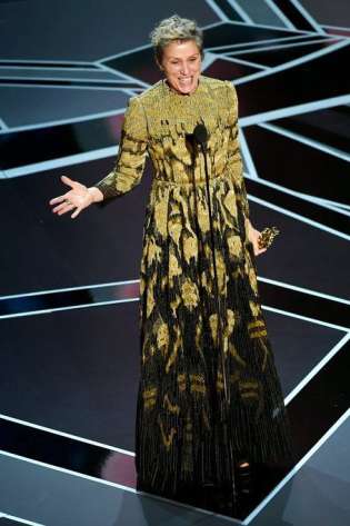 A person in an ornate, long dress stands smiling, gesturing with one hand, holding an award in the other, on a geometrically patterned stage under bright lights.