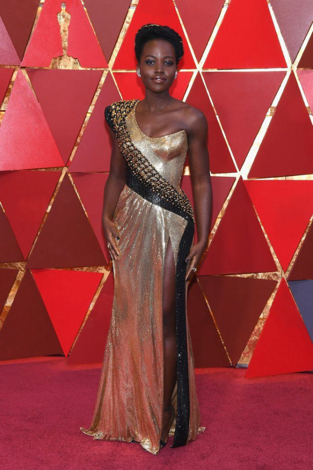 A person stands in a gold gown with black accents and a high slit, posing on a red carpet