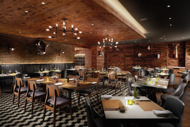 Restaurant features wooden tables and leather chairs set for dining, surrounded by brick and wood walls adorned with mounted animal heads. Warm lighting creates a cozy, rustic atmosphere.