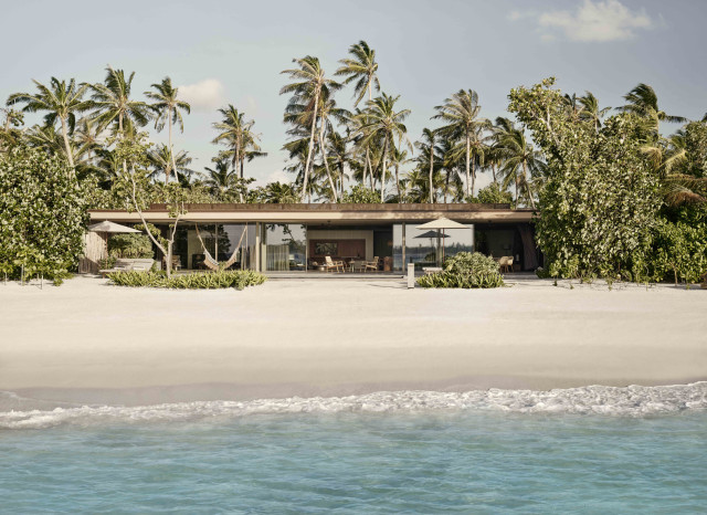 A modern beachfront house with large windows and patio sits quietly among lush palm trees, facing a sandy beach with gentle ocean waves under a partly cloudy sky.