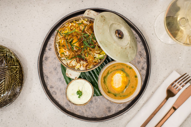 A lidded bowl of biryani and a small bowl of creamy curry rest on a textured round plate, accompanied by yogurt sauce
