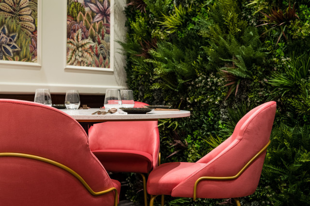 Pink chairs surround a round table set with glasses and cutlery, positioned against a lush green plant wall, with botanical art hung on the adjacent wall