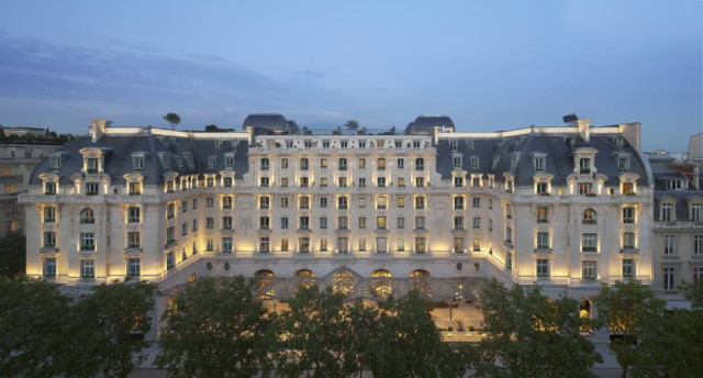 A large, illuminated historic building with mansard roofs stands symmetrically against a twilight sky