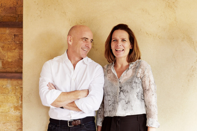 Two people stand side by side, smiling, in front of a beige wall