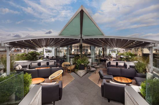 A modern rooftop lounge with black couches and wooden tables is arranged under a triangular canopy, surrounded by glass railings and green plants, overlooking a cityscape