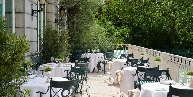 White tablecloth-covered tables with green chairs are arranged on an outdoor patio, surrounded by leafy plants and a beige stone building with ornate wall lamps