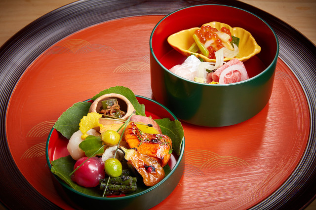 Two green lacquerware bowls display an assortment of vibrant Japanese cuisine, with vegetables, fish, and roe elegantly arranged. The bowls rest on a decorative red and black lacquer tray.