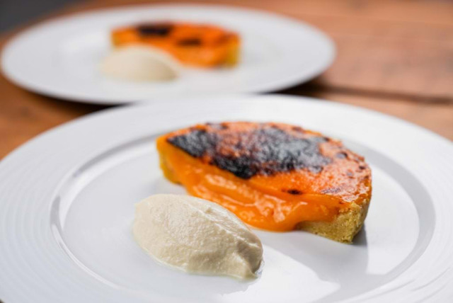 A slice of toasted orange tart melts slightly on a plate beside a dollop of cream; a similar dessert is blurred in the background on a wooden table
