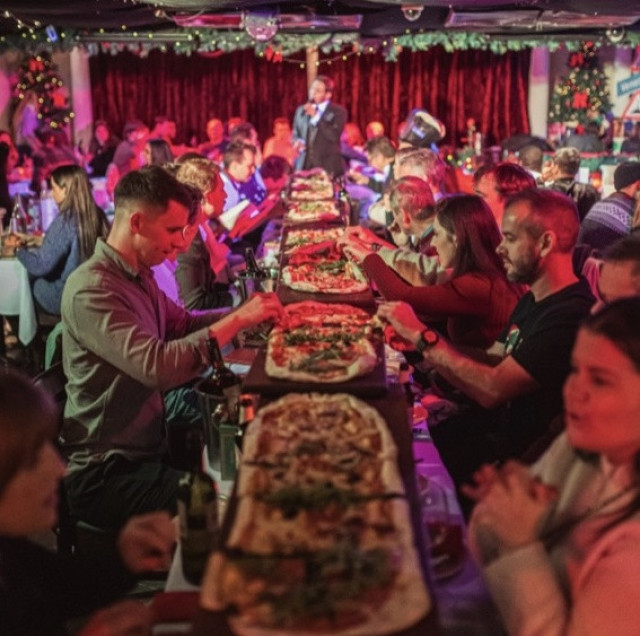 A long table is covered with flatbread pizzas. People seated on both sides are dining in a warmly lit environment, with festive decorations and a performer in the background.
