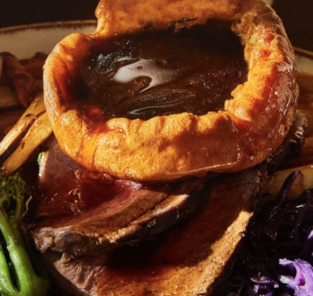 Yorkshire pudding filled with gravy sits atop roast beef slices, surrounded by roasted vegetables and greenery on a plate, creating a hearty, appetizing meal scene.
