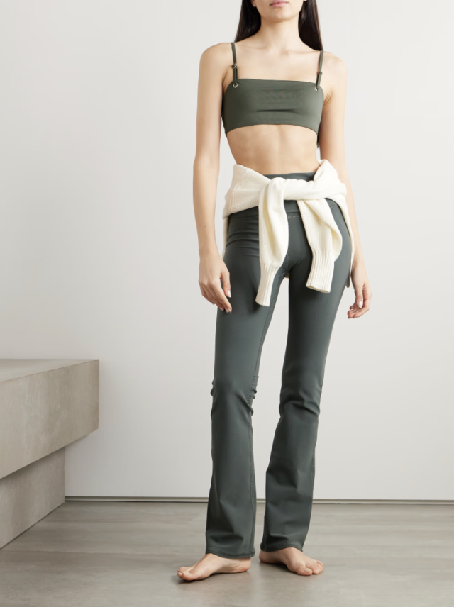 A person stands barefoot in a minimalist room, wearing a green crop top and pants, with a white sweater tied around their waist.