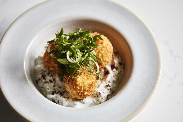Three golden, crispy croquettes rest atop creamy sauce, garnished with fresh green herbs, in a white bowl on a light marble surface