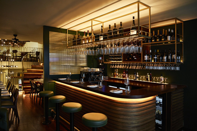 A polished wooden bar counter is lined with stools, displaying wine glasses and bottles overhead